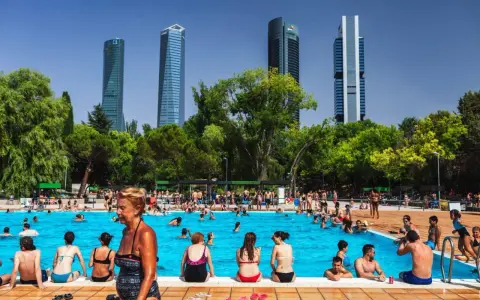 Gente disfrutando en una vibrante piscina comunitaria, con rascacielos modernos de fondo y árboles frondosos.