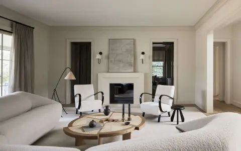 Sofá blanco texturizado, sillones blancos con brazos oscuros, mesa de centro de madera y ratán, chimenea y arte abstracto. Colores que combinan con el blanco crean un ambiente sereno.