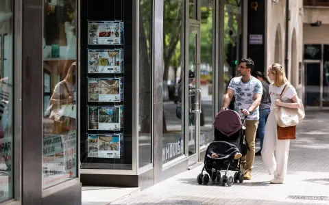 Pareja pasea con carrito, mirando escaparate inmobiliario. Quizás planean donar un piso a un hijo en Cataluña.