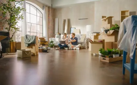 Pareja celebrando mientras planean comprar un piso a medias. Cajas y plantas por doquier, listos para empezar una nueva vida.