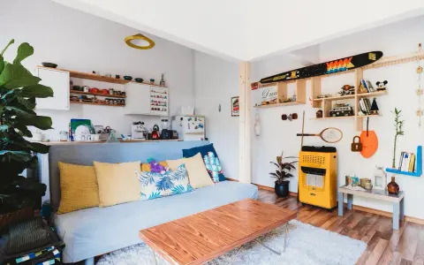 Sala de estar lista para alquilar a estudiantes. Sofá azul, mesa de centro de madera, estanterías con objetos decorativos y cocina pequeña.