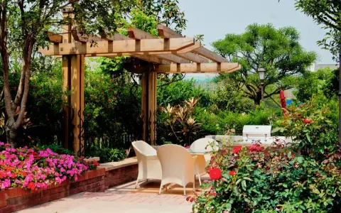 Jardín con pérgola de madera, mesa y sillas, flores rosas y rojas, y barbacoa.