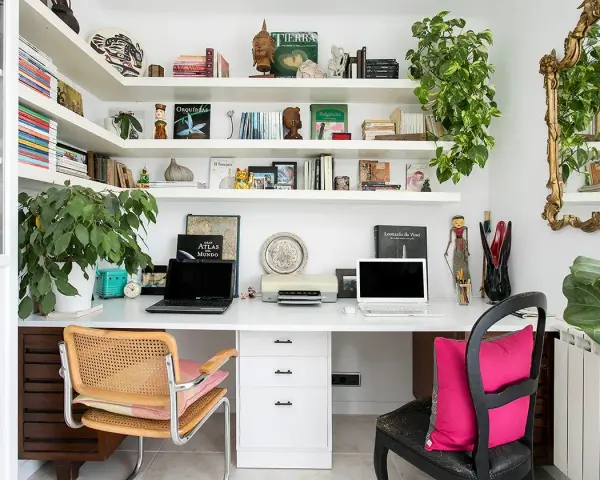 Un despacho en casa con estanterías llenas de libros y plantas. Dos portátiles, una impresora y decoración variada crean un espacio de trabajo funcional y acogedor.