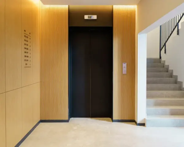 Portal moderno con ascensor negro y escaleras de mármol. Ideas para como decorar un portal de vecinos con paneles de madera y luz cálida.
