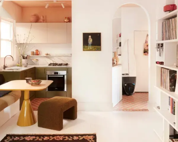 Un salón-cocina acogedor con estanterías llenas de objetos, un sofá cama y una mesa amarilla. Un pasillo arqueado conduce a otra estancia con una tabla de surf. Estas casas curiosas invitan a la vida.