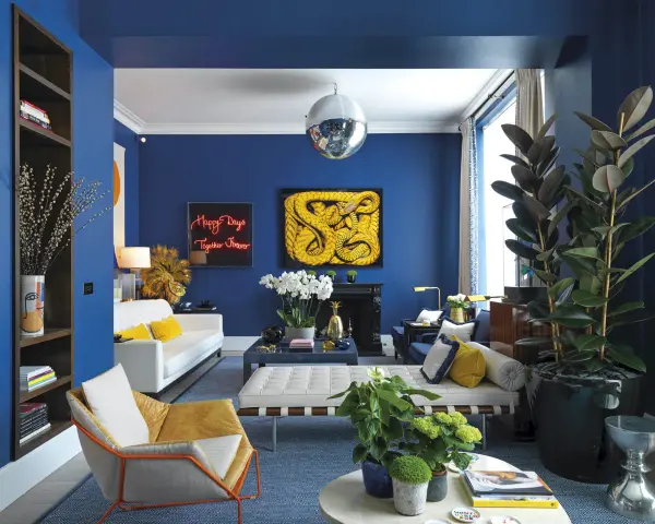 Salón moderno con paredes azul cobalto. Sofá blanco, sillón mostaza y cojines amarillos. Arte abstracto y plantas. Colores que combinan con el azul resaltan.