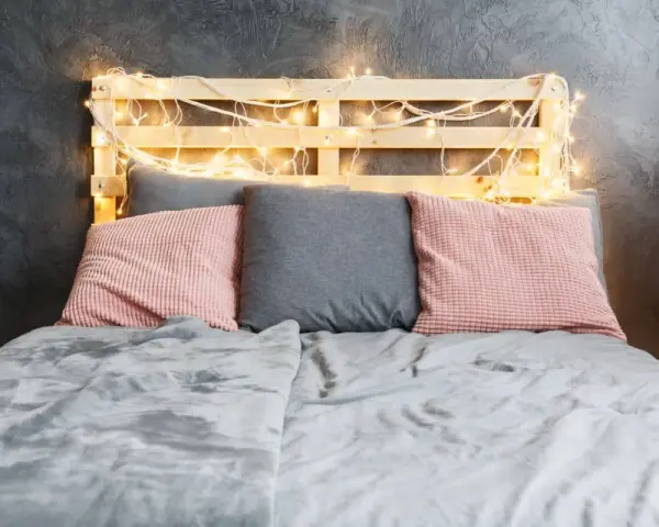 Cabecero de palets ideas: cama con cojines rosas y grises, manta gris y luces cálidas.