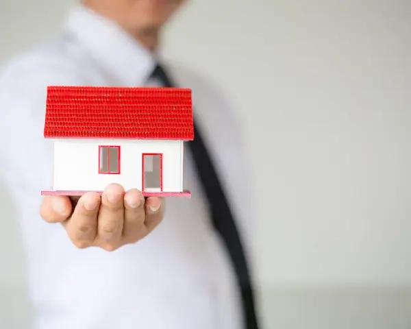 Un hombre con corbata negra sostiene una maqueta de casa blanca con techo rojo. ¿Listo para que puedas alquilar mi vivienda habitual?