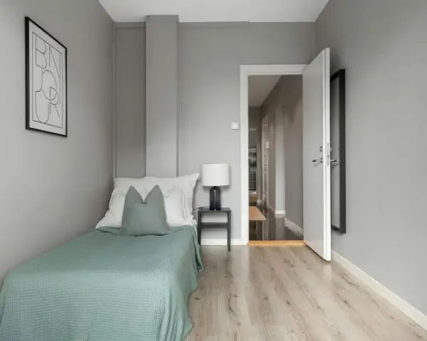 Habitación minimalista con cama individual, mesita de noche y lámpara. La puerta abierta revela un pasillo, sugiriendo que es legal alquilar habitaciones.