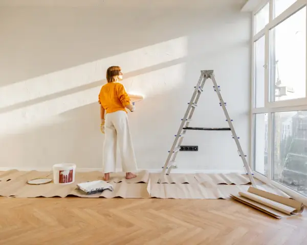 Mujer pintando pared con rodillo, lista para decorar piso antiguo de alquiler. Escalera, cubeta de pintura y protectores de suelo.