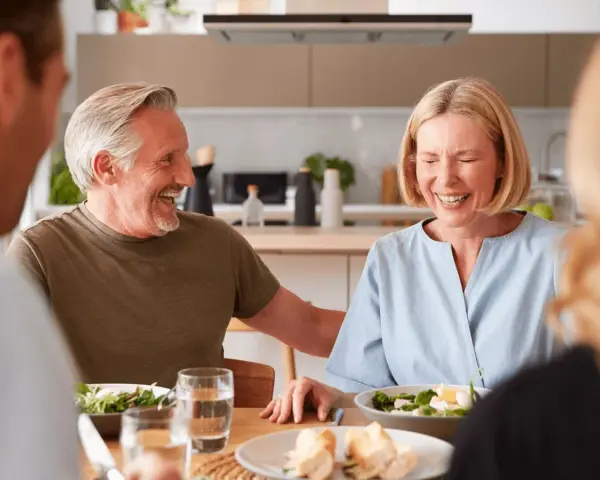 Padre e hijo ríen mientras planean comprar vivienda a medias. Comida familiar en la cocina.