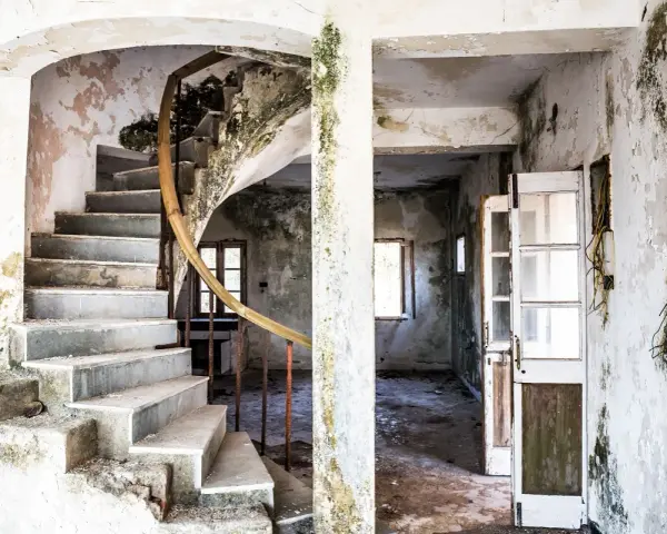 Interior de una casa en ruinas con escalera de caracol y paredes desconchadas. El estado de abandono evidencia las obligaciones del ayuntamiento para su mantenimiento.