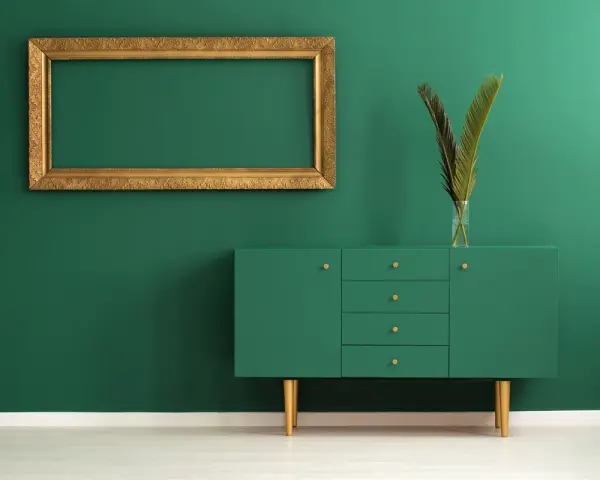 Un mueble verde esmeralda con patas doradas y un marco dorado vacío sobre pared verde. Los colores que pegan con el dorado son el verde intenso y el blanco.