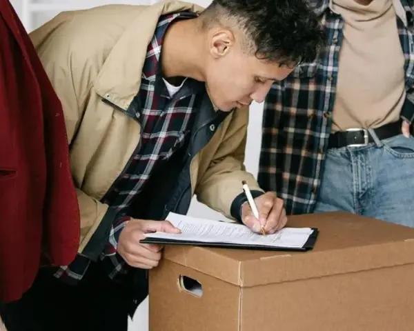 Joven firma un contrato de alquiler sobre una caja de mudanza, con otras dos personas observando.