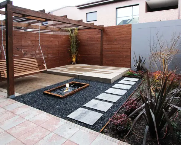 Un columpio de madera cuelga bajo un cenador. Senderos de piedra cruzan grava negra hacia un estanque. Minimalistas jardines modernos.
