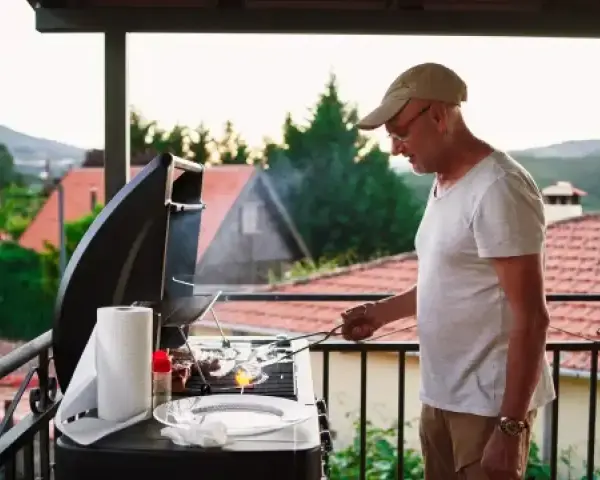 Hombre mayor cocina en barbacoa en terraza. ¡Cuidado con las multas por hacer barbacoas en terrazas!