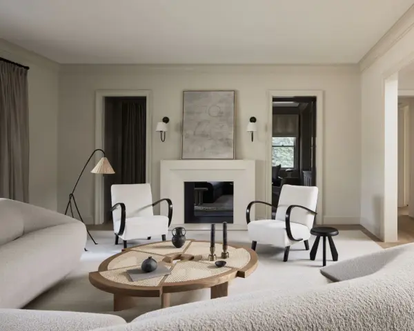 Sofá blanco texturizado, sillones blancos con brazos oscuros, mesa de centro de madera y ratán, chimenea y arte abstracto. Colores que combinan con el blanco crean un ambiente sereno.