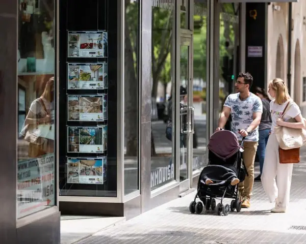 Pareja pasea con carrito, mirando escaparate inmobiliario. Quizás planean donar un piso a un hijo en Cataluña.