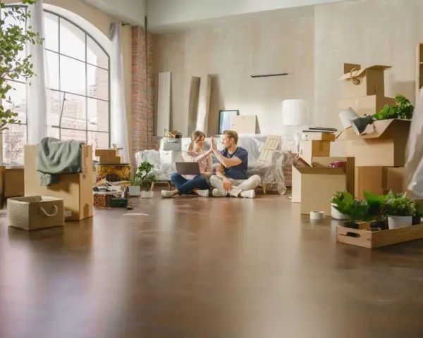 Pareja celebrando mientras planean comprar un piso a medias. Cajas y plantas por doquier, listos para empezar una nueva vida.