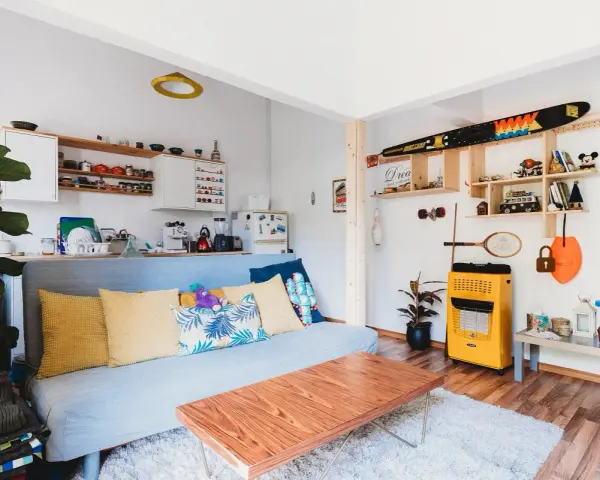 Sala de estar lista para alquilar a estudiantes. Sofá azul, mesa de centro de madera, estanterías con objetos decorativos y cocina pequeña.