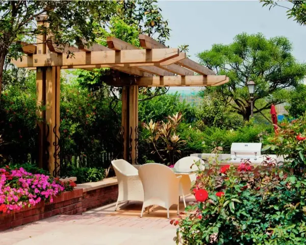 Jardín con pérgola de madera, mesa y sillas, flores rosas y rojas, y barbacoa.