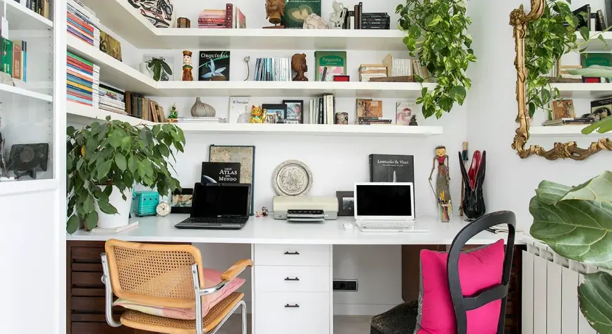Un despacho en casa con estanterías llenas de libros y plantas. Dos portátiles, una impresora y decoración variada crean un espacio de trabajo funcional y acogedor.