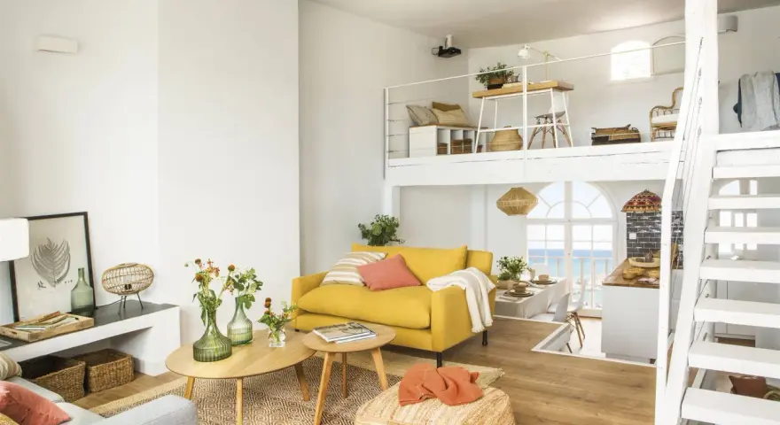 Un salón luminoso con sofá amarillo, mesas de madera y una escalera blanca que lleva a un altillo. Parece uno de esos **casas de cuento** con vistas al mar.