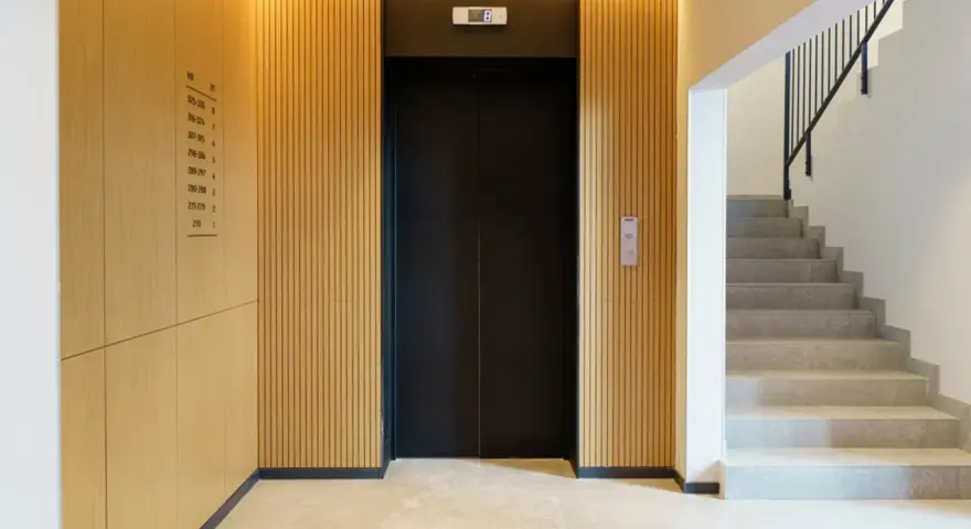 Portal moderno con ascensor negro y escaleras de mármol. Ideas para como decorar un portal de vecinos con paneles de madera y luz cálida.