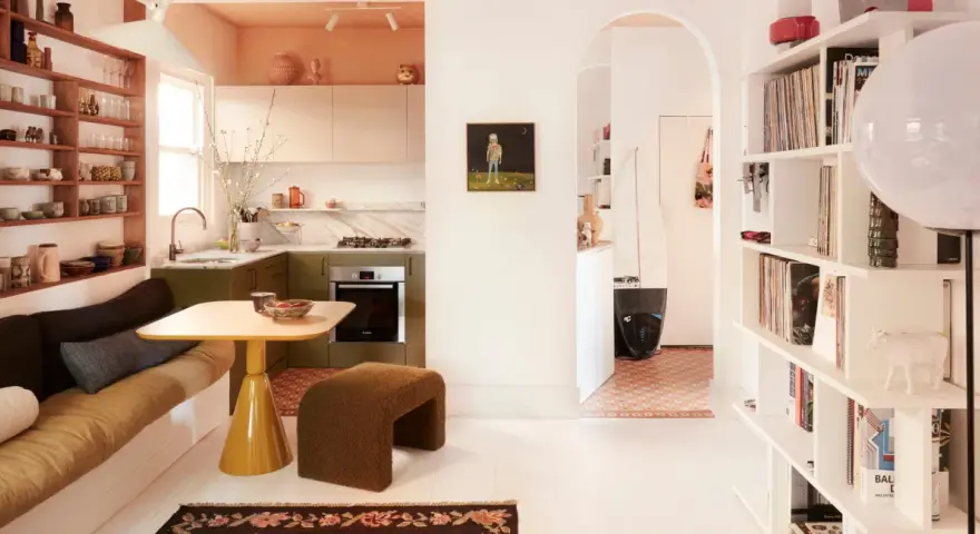 Un salón-cocina acogedor con estanterías llenas de objetos, un sofá cama y una mesa amarilla. Un pasillo arqueado conduce a otra estancia con una tabla de surf. Estas casas curiosas invitan a la vida.