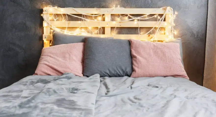 Cabecero de palets ideas: cama con cojines rosas y grises, manta gris y luces cálidas.
