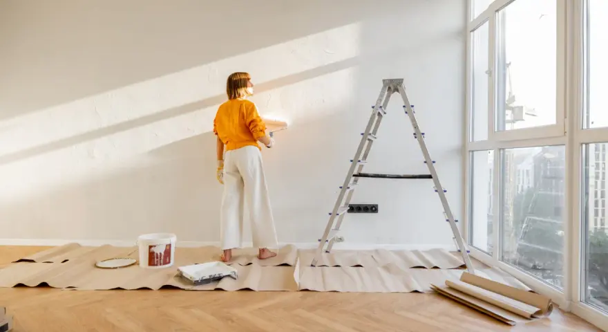 Mujer pintando pared con rodillo, lista para decorar piso antiguo de alquiler. Escalera, cubeta de pintura y protectores de suelo.