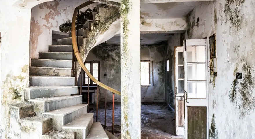 Interior de una casa en ruinas con escalera de caracol y paredes desconchadas. El estado de abandono evidencia las obligaciones del ayuntamiento para su mantenimiento.