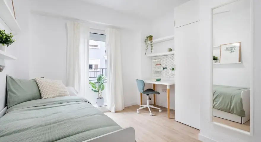 Habitación de estudiante con cama, escritorio, silla y espejo.