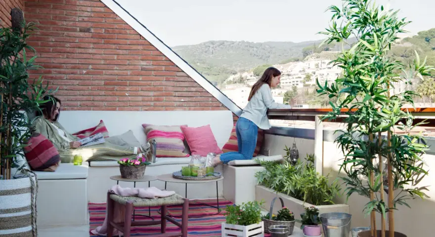 Dos mujeres disfrutan de una terraza con arboles bonitos, cojines coloridos y vistas a la montaña.