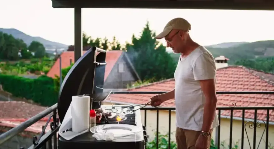 Hombre mayor cocina en barbacoa en terraza. ¡Cuidado con las multas por hacer barbacoas en terrazas!