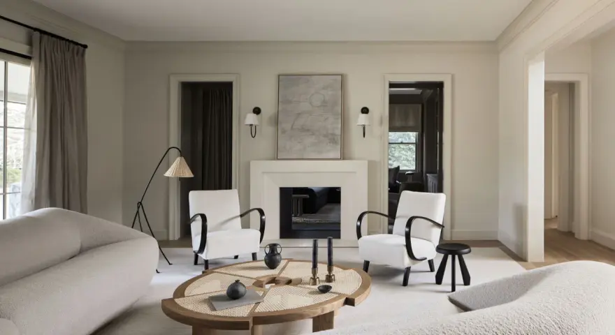 Sofá blanco texturizado, sillones blancos con brazos oscuros, mesa de centro de madera y ratán, chimenea y arte abstracto. Colores que combinan con el blanco crean un ambiente sereno.