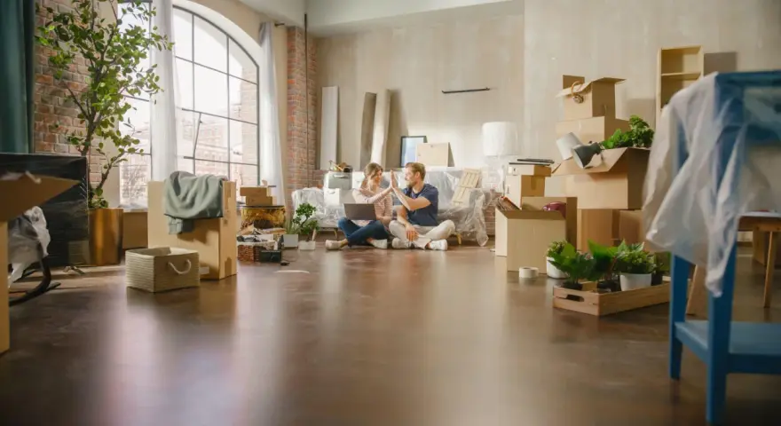 Pareja celebrando mientras planean comprar un piso a medias. Cajas y plantas por doquier, listos para empezar una nueva vida.
