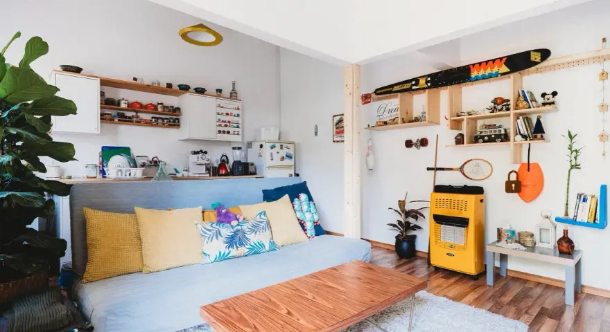 Sala de estar lista para alquilar a estudiantes. Sofá azul, mesa de centro de madera, estanterías con objetos decorativos y cocina pequeña.