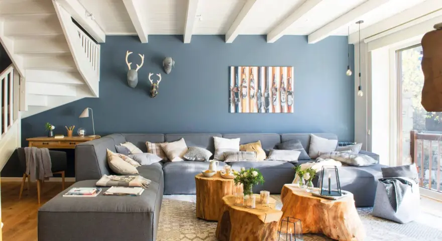 Sofá modular gris y pared azul crean un ambiente acogedor. Mesas de tronco y detalles en madera aportan calidez.