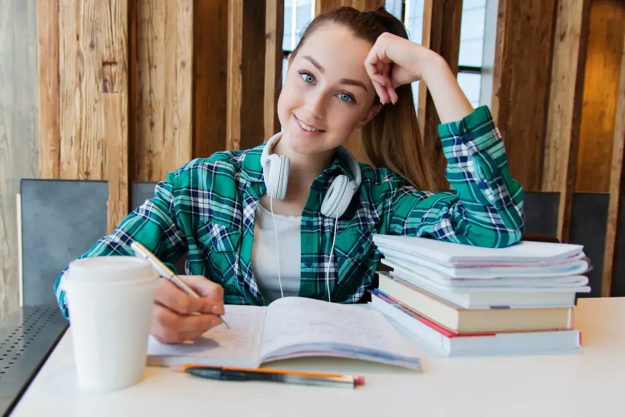 Estudiante sonriente con auriculares, escribiendo en un cuaderno. A su lado, una pila de libros y un café. Ideal para un contrato de arrendamiento de habitaciones para estudiantes.