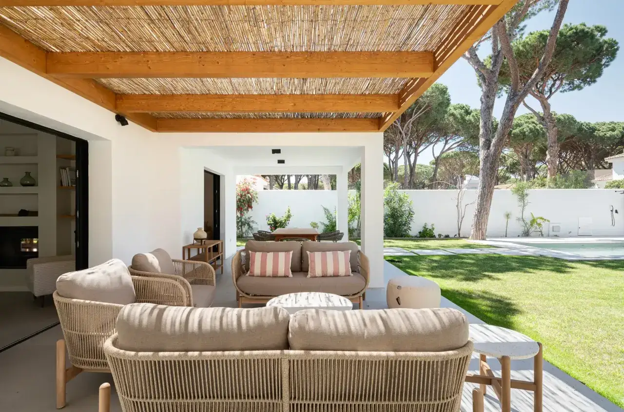 Terraza con muebles de rat&aacute;n y cojines, ideal para decorar casa playa poco presupuesto. P&eacute;rgola de ca&ntilde;a y madera, jard&iacute;n verde y piscina al fondo.