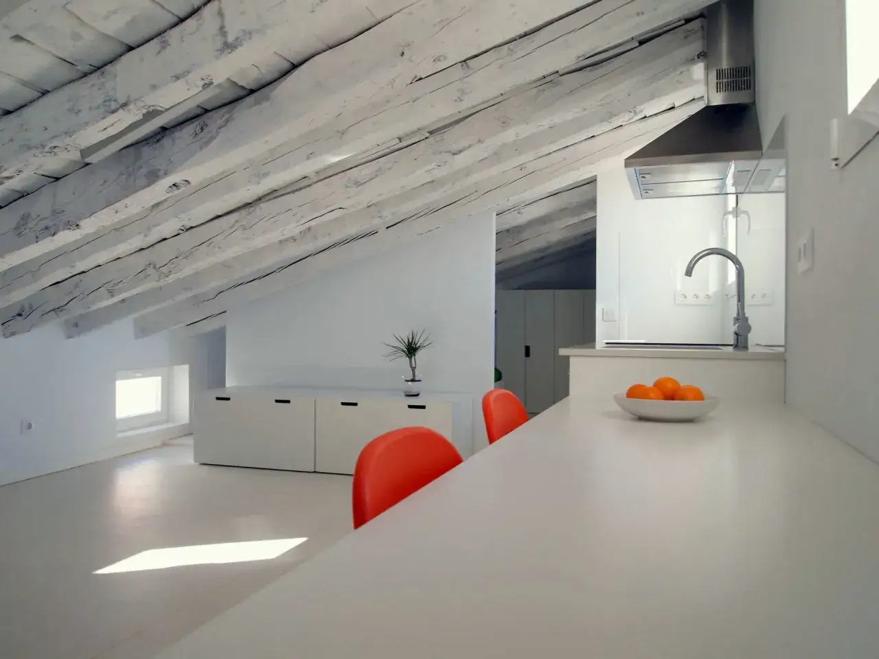 Cocina moderna bajo un techo de vigas de madera, creando una buhardilla con encanto. Dos sillas naranjas destacan en el espacio.