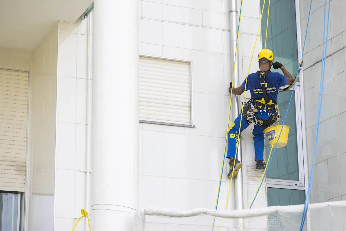 Trabajador en altura, con arn&eacute;s y casco amarillo, desciende por la fachada de un ite de edificios blancos, con un cubo amarillo colgando.