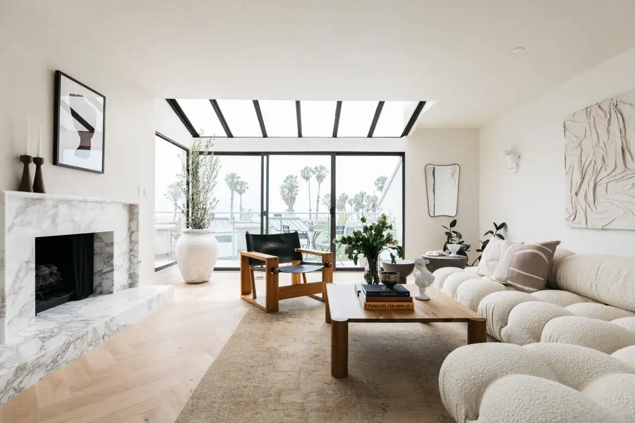 Salones blancos y madera con chimenea de m&aacute;rmol, sof&aacute; texturizado y vistas a palmeras.