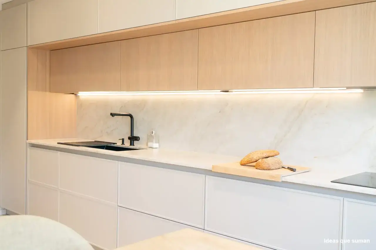 Cocina blanca y gris claro con encimera de mármol, grifo negro y pan en tabla de cortar.