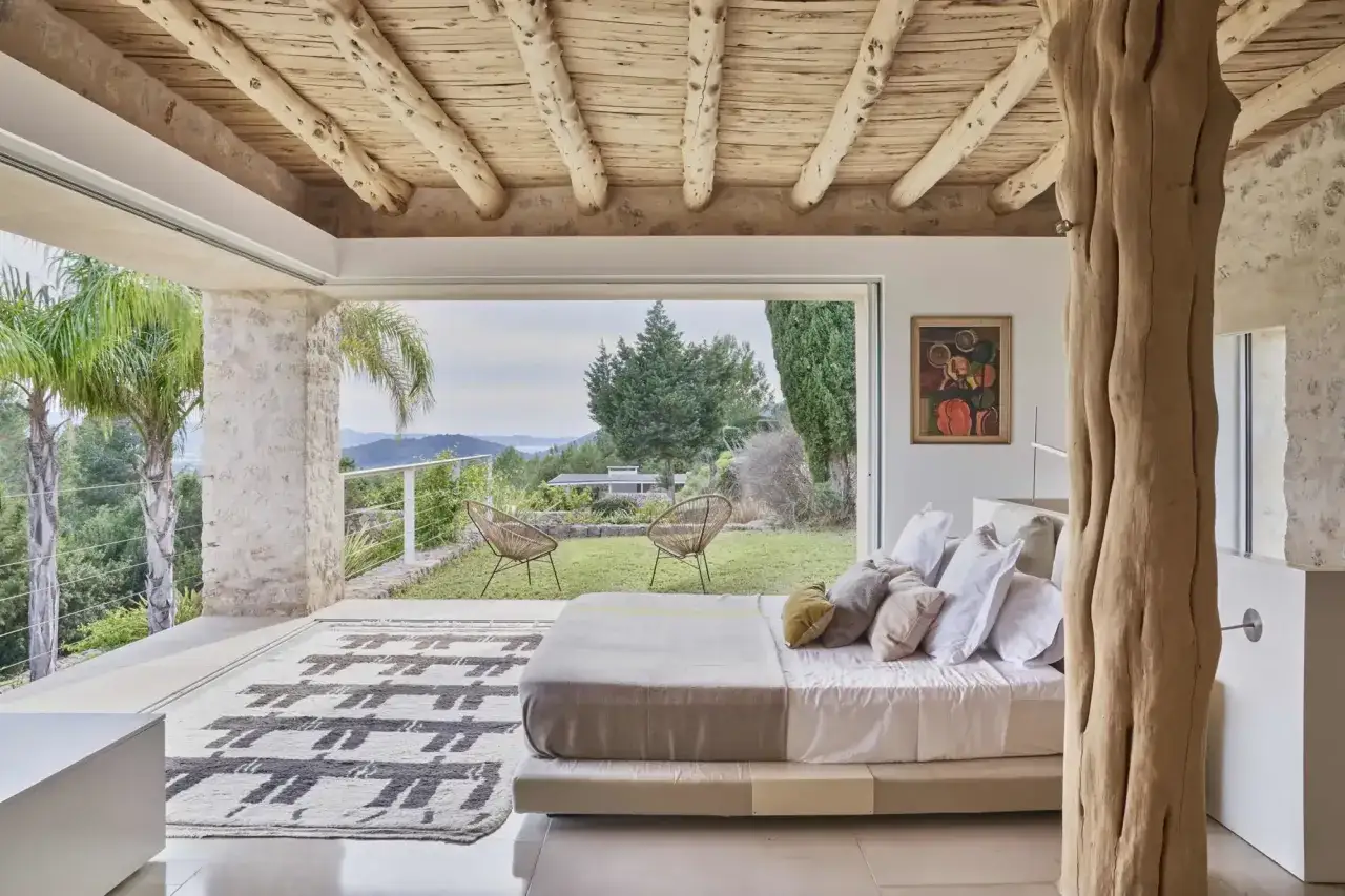 Dormitorio con cama, alfombra y vistas al exterior. La decoraci&oacute;n ibicenca exterior se fusiona con el interior r&uacute;stico.