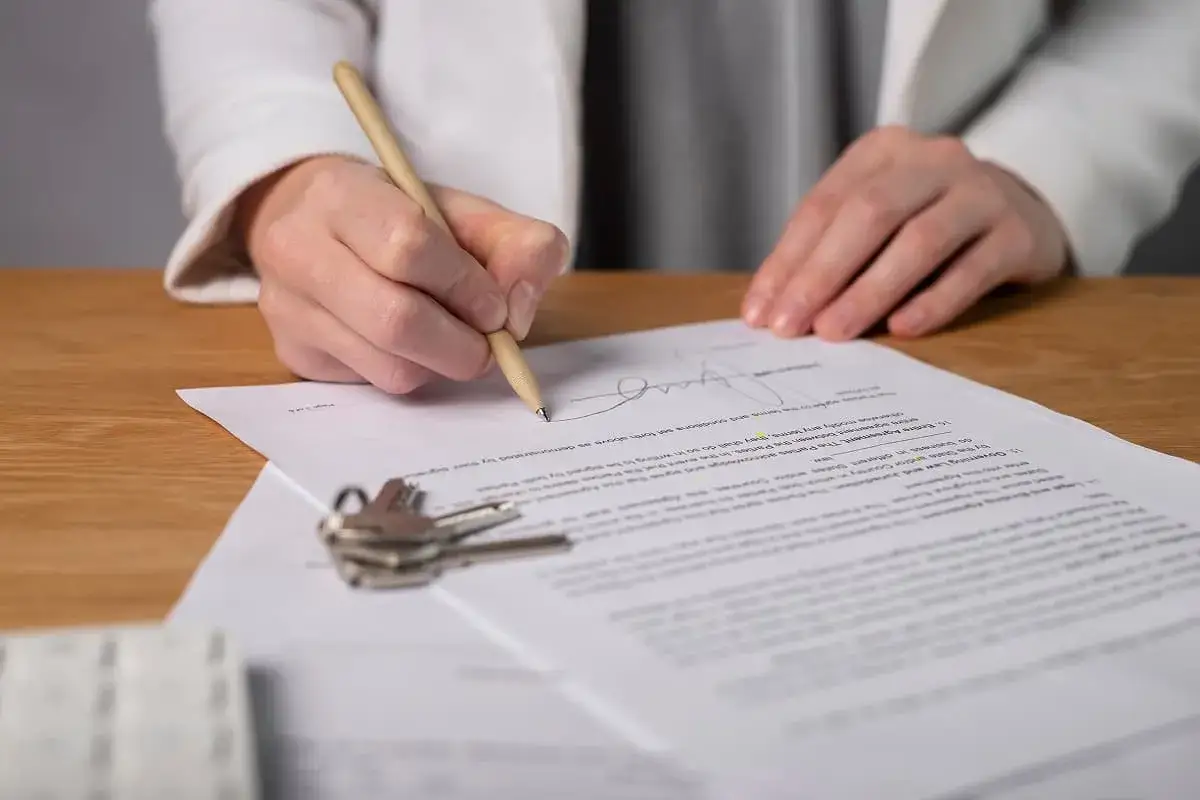 Persona firma un documento, que es la escritura de una casa, con un bol&iacute;grafo. Llaves y papeles sobre la mesa.