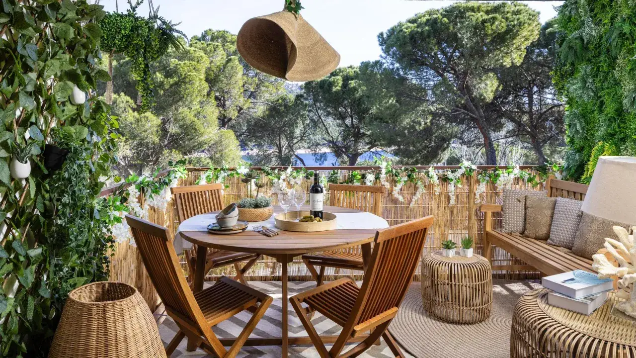Un jard&iacute;n bonito en un balc&oacute;n con mesa de madera, sillas, banco y mucha vegetaci&oacute;n.