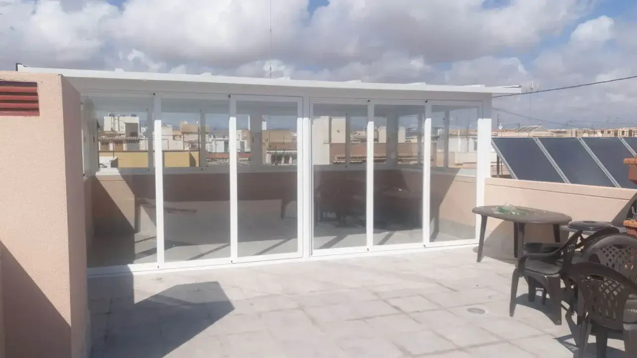 Techar patio comunitario de uso privativo con cerramiento de cristal y aluminio. Muebles de exterior y paneles solares al fondo.