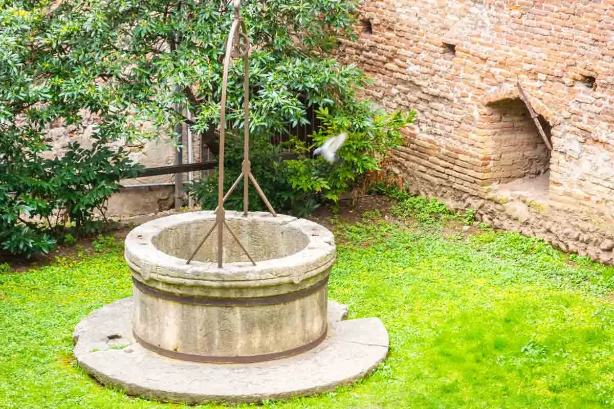 Pozo antiguo de piedra con estructura metálica, rodeado de césped y vegetación, junto a un muro de ladrillos. Ideal para quienes buscan cómo legalizar un pozo ya hecho.